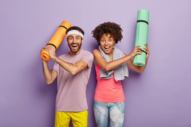 运动装快乐的女人和男人在有氧运动后开心的水平镜头 用折叠的卡雷马特举手 穿着运动服 享受运动的业余时间 孤立在紫色的墙上多样化的夫妇运动员高兴男性