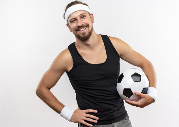 腕带面带微笑的年轻帅气的运动型男人戴着头带和腕带 手放在腰上拿着足球 隔离在白色的空间里英俊男人戴着