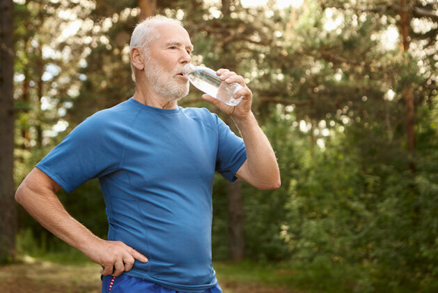 活动照片中是一位富有魅力的男性退休老人 秃头 留着胡茬 站在松林旁 手里拿着一瓶饮用水 在户外慢跑后 神清气爽喝水户外退休