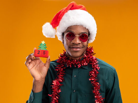 花环戴着圣诞帽 戴着花环 戴着太阳镜的非裔美国人拿着玩具方块 站在橙色的墙上微笑着和25岁约会日期圣诞节非洲