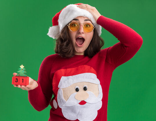 站着穿着圣诞毛衣 戴着圣诞帽 戴着圣诞眼镜 手里拿着新年礼物的玩具立方体的小女孩站在绿色的墙边 惊讶不已日期惊讶圣诞老人