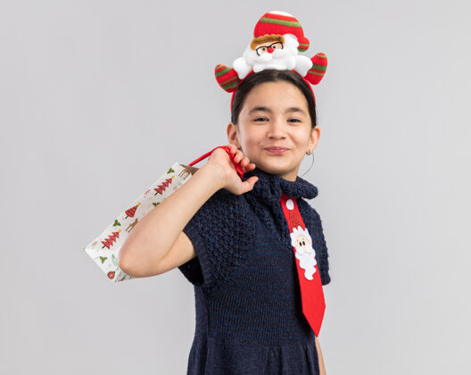积极小女孩穿着针织连衣裙 头上系着一条红色领带 头上戴着一个有趣的圣诞镶边 手里拿着一个装有圣诞礼物的纸袋 看上去快乐而积极年新年看