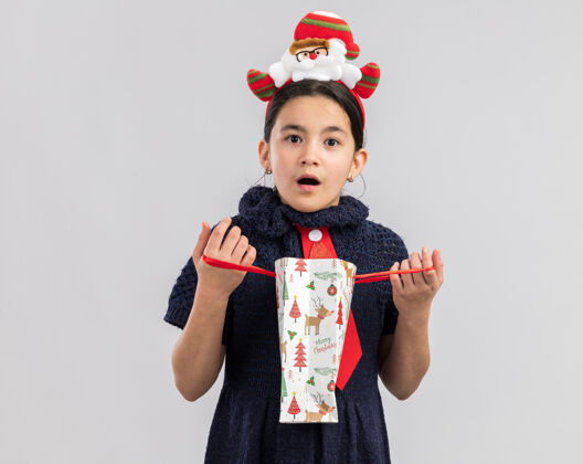 新年小女孩穿着针织连衣裙 头上系着一条红色领带 带着有趣的圣诞边 打开一个装有圣诞礼物的纸袋 看上去很惊讶领带年惊讶