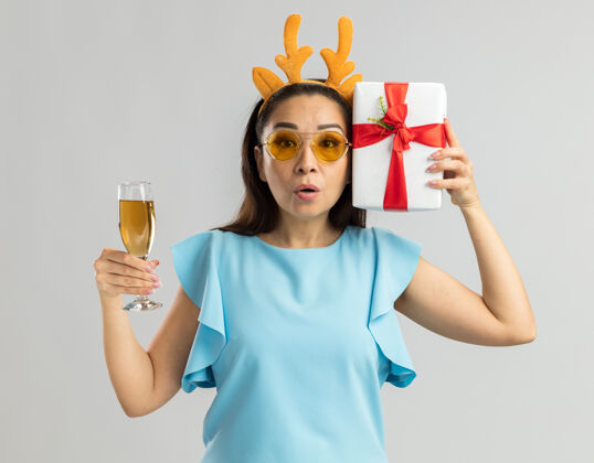 圣诞身穿蓝色上衣的年轻女子戴着滑稽的鹿角边 戴着黄色眼镜 手里拿着一杯香槟和圣诞礼物 看上去既惊讶又担心女人年举着