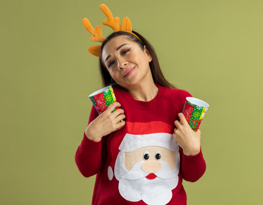 新穿着红色圣诞毛衣的年轻女子 戴着滑稽的鹿角边 手里拿着五颜六色的纸杯 脸上洋溢着积极的情绪 面带微笑鹿站着穿着