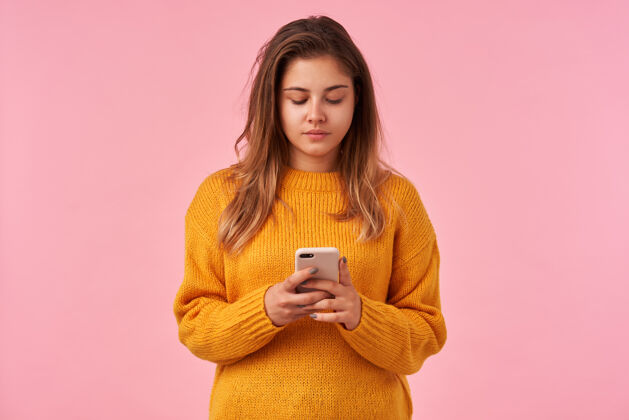 摆姿势苏迪奥的画像 年轻的棕色头发的女士 自然化妆手持智能手机 而站在粉红色的 注视着屏幕 并保持她的嘴唇折叠智能手机羊毛衫衣服