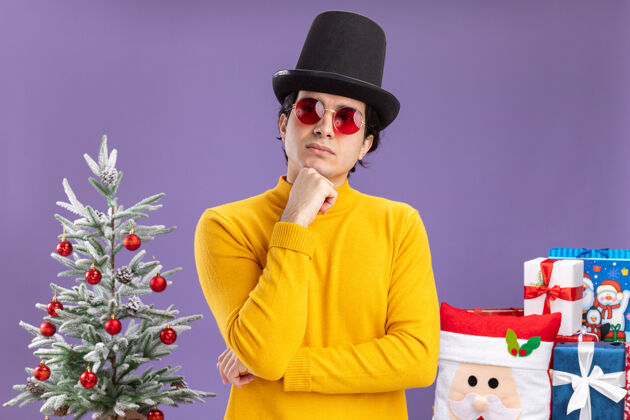 思考一个穿着黄色高领毛衣戴着黑色帽子的年轻人 手放在下巴上 一边看一边想着站在一棵圣诞树旁边 在紫色背景下送礼物树手帽子