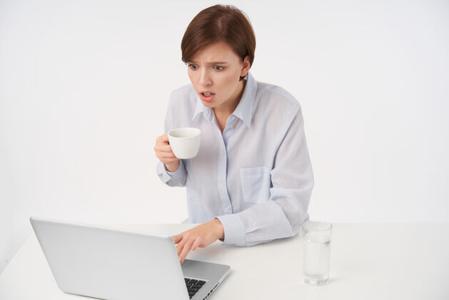 优雅年轻漂亮的棕色头发的女人睁着眼睛 留着随意的发型 手里拿着陶瓷杯 惊奇地看着笔记本电脑的屏幕 读着意想不到的新闻 孤零零地站在白色的地板上咖啡工作自然
