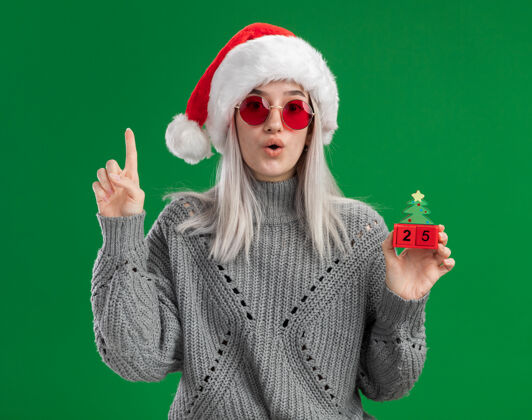 惊讶穿着冬季毛衣 戴着圣诞帽 戴着红眼镜的金发女郎拿着玩具立方体 圣诞节的日期看起来很惊讶 食指站在绿色的背景上索引手指毛衣