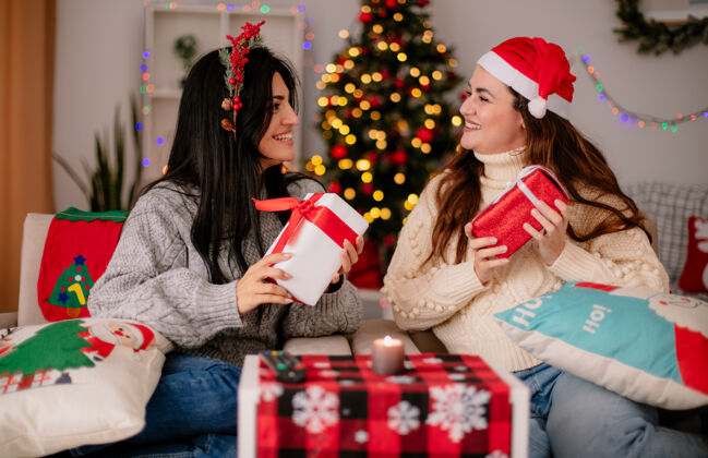 盒子带着圣诞帽和冬青花环的年轻女孩们微笑着拿着礼盒 坐在扶手椅上互相看着 在家里享受圣诞节时光坐着每个时间