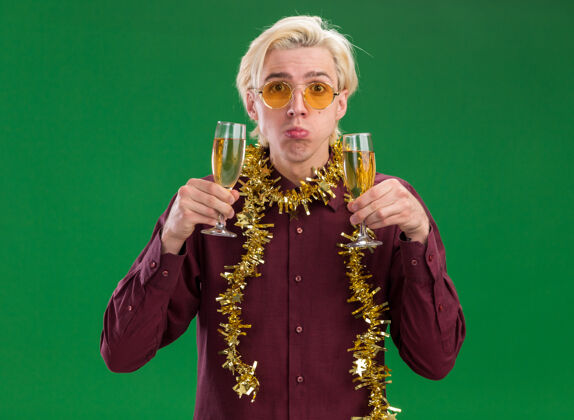 脖子笨手笨脚的年轻金发男子戴着一副戴着金箔花环的眼镜 手里拿着两杯香槟 隔离在绿色的墙上男人圣诞节金发