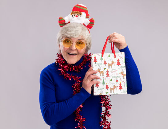圣诞戴着太阳眼镜 脖子上戴着圣诞老人头带和花环 手里拿着纸质礼品袋的惊讶的老妇人花环抱着惊讶