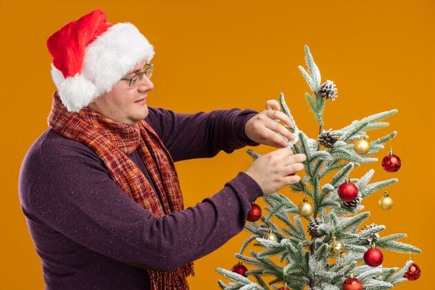 帽子戴着眼镜 戴着圣诞帽 脖子上围着围巾的成年男子站在圣诞树旁的侧视图中 将圣诞树装饰在橙色的背景上圣诞快乐站立周围