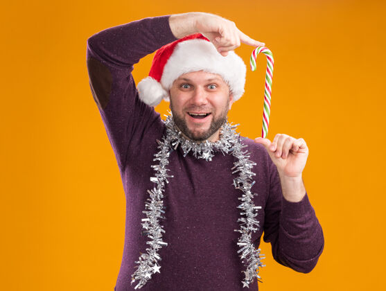 手杖快乐的中年男子戴着圣诞帽 脖子上戴着金箔花环 手持圣诞甜手杖 靠近隔离在橙色墙上的头圣诞老人花环脖子