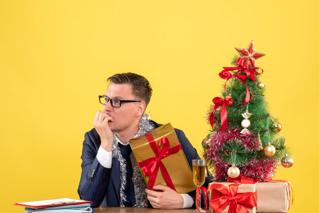 电话前视图困惑的人看着左边坐在圣诞树旁的桌子上 黄色的礼物困惑男人成年人