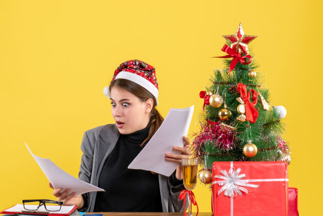 庆典前视图困惑的女孩戴着圣诞帽坐在桌旁看着圣诞树和礼物的文件鸡尾酒鸡尾酒节日女孩