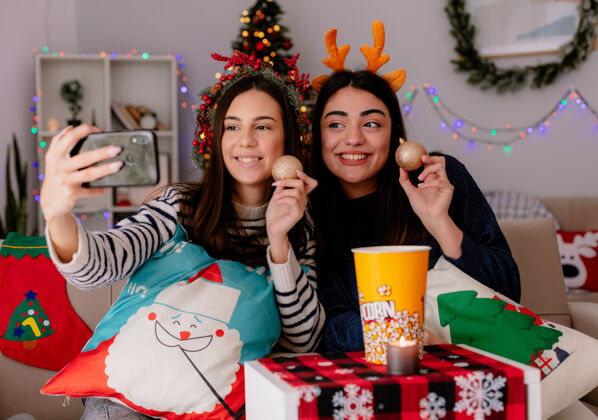 头带带着冬青花环和驯鹿头带的年轻女孩们微笑着拿着玻璃球饰品 坐在扶手椅上自拍 在家里享受圣诞节时光微笑自拍家
