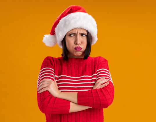 圣诞节闷闷不乐的年轻女孩戴着圣诞帽 站在那里 闭着身子 看着橘色墙壁上孤立着的侧面鼓鼓囊囊的脸颊 留着复制空间帽子站立脸颊