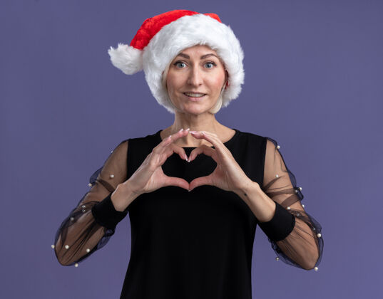 微笑微笑的中年金发女人戴着圣诞帽看着镜头做着心形标志孤立在紫色背景上相机中年帽子