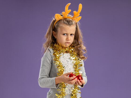 头发悲伤的小女孩戴着圣诞发箍 脖子上戴着花环 手里拿着蓝色背景上孤立的圣诞球圣诞节女孩小