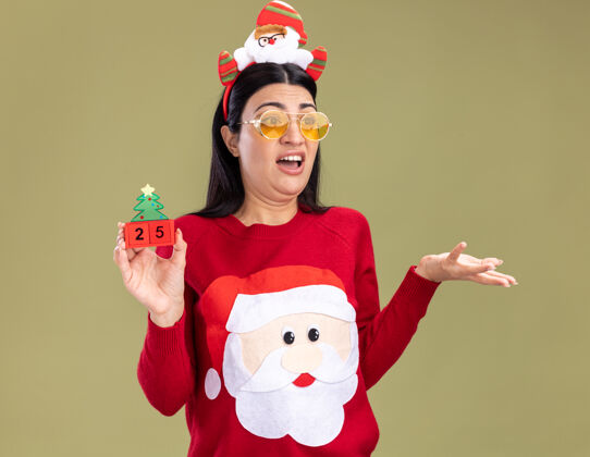 圣诞老人恼怒的年轻白人女孩戴着圣诞老人的头带和毛衣戴着眼镜拿着圣诞树玩具和日期看一边显示空手孤立在橄榄绿的墙上烦恼显示年轻