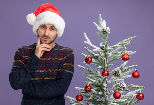 相机体贴的年轻白种人戴着圣诞帽站在装饰好的圣诞树旁 手放在下巴上 看着紫色背景上孤立的摄像机看保持装饰