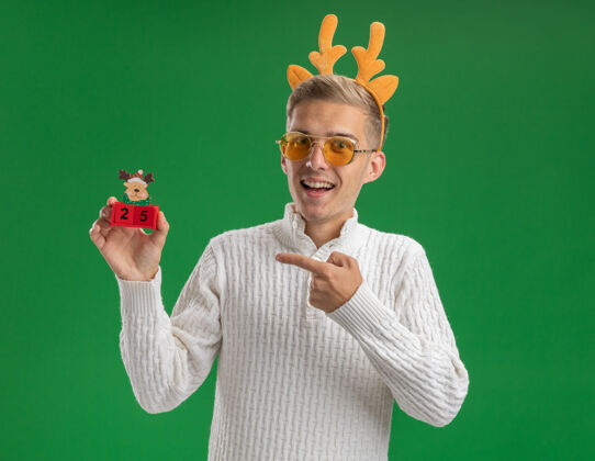 印象深刻印象深刻的年轻帅哥戴着驯鹿鹿角头带戴着眼镜拿着圣诞树玩具和日期隔离在绿色墙上驯鹿日期帅气