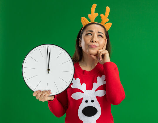 年轻穿着红色圣诞毛衣的年轻女子 戴着滑稽的鹿角边 手持挂钟 站在绿色的背景上困惑地仰望着有趣边缘拿着