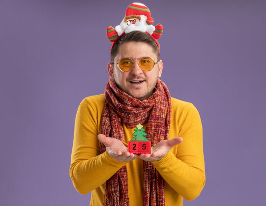 二十快乐的年轻人穿着黄色高领毛衣 戴着暖和的围巾和眼镜 戴着滑稽的帽沿 头上戴着圣诞老人 在紫色的背景上展示着25号玩具立方体圣诞老人头温暖