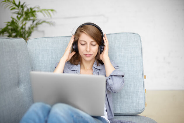 音乐现代科技 乐趣和放松的概念漂亮的女人在室内度过美好的时光 通过无线耳机听音乐 坐在沙发上 浏览网站 搜索新歌漂亮房子微笑