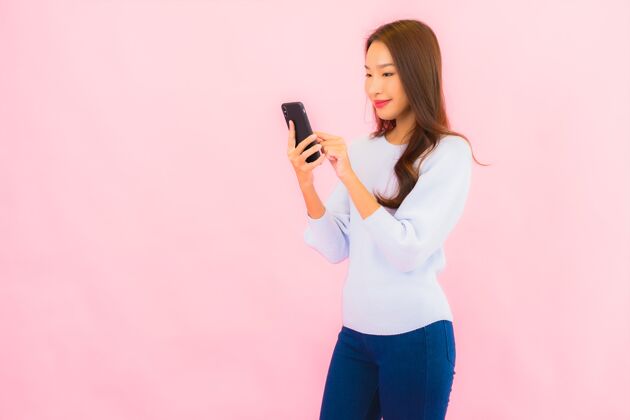 聪明肖像美丽的亚洲年轻女子在粉色隔离墙上使用智能手机短信多彩指着