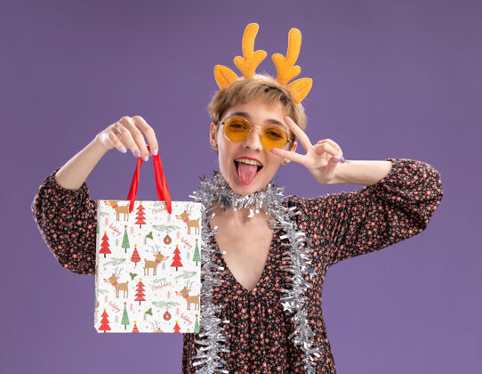 和平俏皮的年轻漂亮女人戴着驯鹿鹿角头带 脖子上戴着金属丝花环 戴着眼镜 手里拿着圣诞礼品袋眼镜驯鹿包