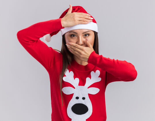 嘴高兴的年轻亚洲女孩戴着圣诞帽与毛衣覆盖额头和嘴与双手隔离在白色背景毛衣圣诞节女孩