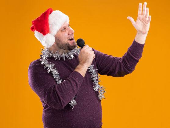 金属片快乐的中年男子戴着圣诞帽 脖子上戴着金箔花环 手持麦克风 抬头举手 在橙色背景下独自歌唱麦克风花环手