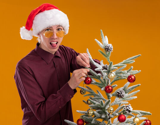 个人资料一个戴着圣诞帽和眼镜的可爱的金发年轻人站在圣诞树旁的侧视图里 用圣诞饰品装饰圣诞树 看着相机 在橙色的背景上露出孤立的舌头舌头金发帽子