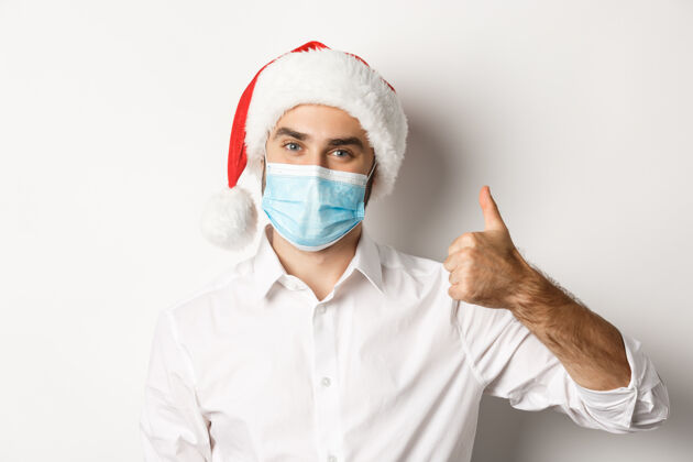 西装戴着面具和圣诞帽的满意男人竖起大拇指 用预防措施庆祝圣诞节满意竖起大拇指蓄胡子