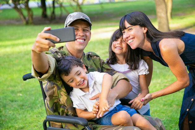 女人快乐快乐的残疾军人和他的妻子和两个孩子在公园里自拍家庭团聚和支持的概念制服手机高加索