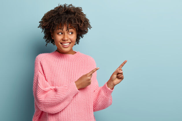 惊奇快乐快乐的女人的肖像在复制空间指向正确的方向 快乐地凝视着 广告好的产品 穿着超大号的粉红色套头衫 隔离在蓝色的墙上看看那里的酷广告促销牙关非洲