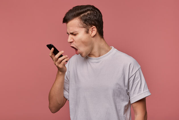 情绪一张室内照片 愤怒的年轻黑发男性手拿手机站在粉色背景上 在电话里说些不愉快的话 并对着手机怒目而视地喊叫年轻听力家伙