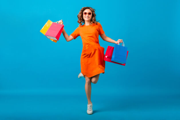 年轻迷人的快乐微笑时尚女性购物狂穿着橙色时髦的超大号连衣裙蹦蹦跳跳拿着蓝色背景的购物袋包商店漂亮