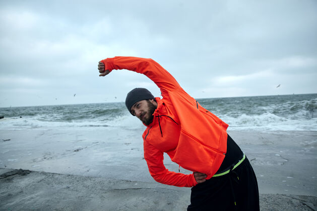 帅气在灰色寒冷的早晨 一位留着胡须的年轻黑发运动型男子在海边做体育锻炼 在锻炼前伸展肌肉健身男模海滩海滨训练