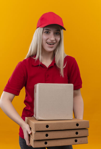 穿着微笑的小女孩穿着红色t恤 戴着帽子 戴着牙套 拿着盒子和披萨盒 背景是孤立的橙色抱着送货T恤