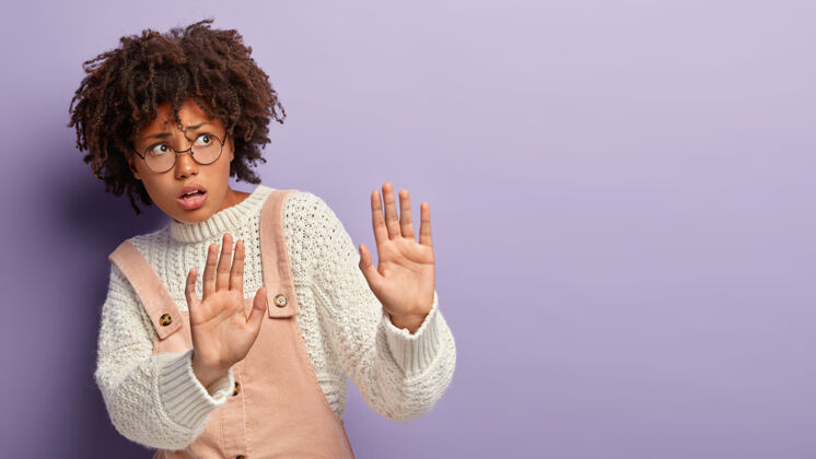 理发惊恐的黑皮肤女性伸出手掌 做出保护的手势 有着担心紧张的表情 要求不要靠近 戴着眼镜和工作服 孤立在紫色的墙上吓坏跳投禁止