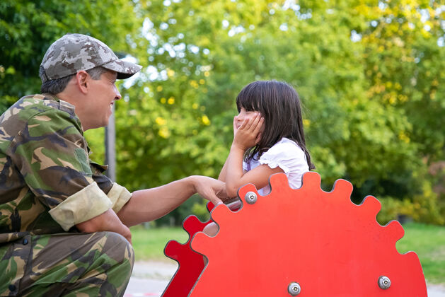 军队快乐的军旅爸爸和小女儿在操场上玩 和小女孩一起聊天和玩耍 小女孩骑着摇摆刺猬做父母还是童年的概念黑色摇摆成人