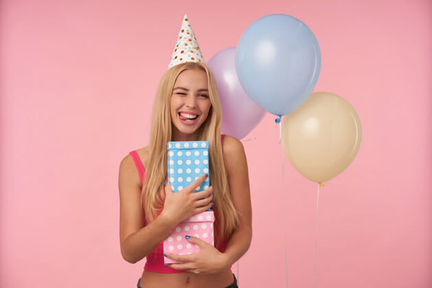 庆祝在室内拍摄的积极年轻的长发女性高兴 而在五彩气球摆姿势 在生日派对上玩得开心 拿着礼物 站在粉红色的背景20多岁室内女性