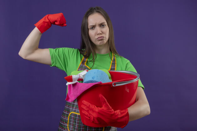 制服悲伤的清洁小女孩穿着制服戴着手套拿着清洁工具在紫色背景上做着强烈的手势紫色清洁手套