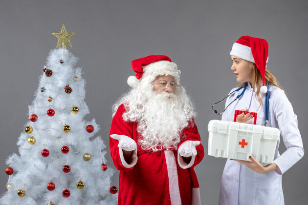 帽子圣诞老人和女医生的前视图 女医生拿着急救箱在灰色的墙上援助Covid节日