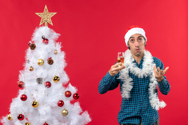 圣诞帽圣诞节心情与关心的年轻人在圣诞树附近举起一杯葡萄酒 圣诞老人帽子在一个蓝色条纹衬衫圣诞老人装饰新年