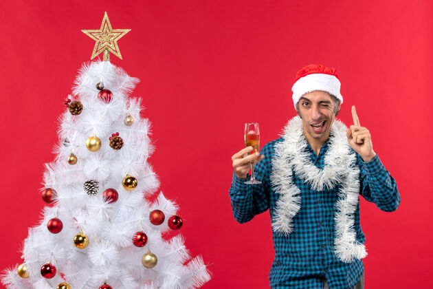 新年前夜圣诞节心情自信快乐疯狂情绪化的年轻人戴着圣诞老人帽穿着蓝色条纹衬衫在圣诞树旁举起一杯葡萄酒圣诞年圣诞树