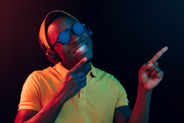 非洲年轻帅气快乐的时髦男人在霓虹灯下用耳机在黑工作室听音乐迪斯科舞厅 夜总会 嘻哈风格 积极的情绪 面部表情 舞蹈概念听音频舞蹈